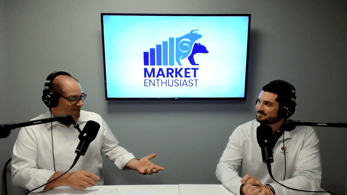two men, wearing white button-down shirts, sit in front of a tv screen while recording a podcast
